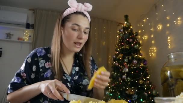 Joven hermosa mujer corta papas para ensalada con un cuchillo para olivier en una tabla de cortar de madera en un ambiente festivo de año nuevo con un árbol de Navidad — Vídeos de Stock