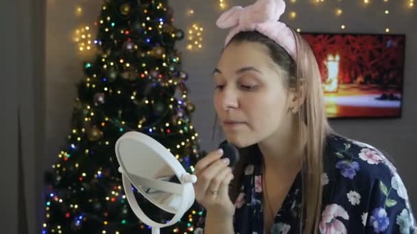 Une femme prend soin de sa belle peau sur son visage, debout près du sapin de Noël. Jeune femme appliquant fond de teint sur le visage avec éponge — Video