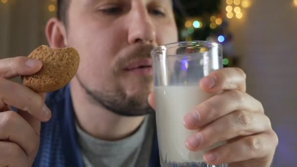 A bald man at night in his apartment eats homemade delicious Christmas gingerbread cookies with a glass of milk near a Christmas tree with a garland — Stock Video