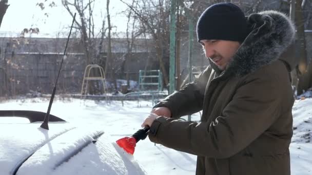 Un uomo con un cappello con un raschietto in inverno pulisce il parabrezza o il parabrezza di un'auto da neve e ghiaccio. Utilizzare un raschietto o raschietto. clima freddo. nel pomeriggio nel parcheggio nel cortile — Video Stock