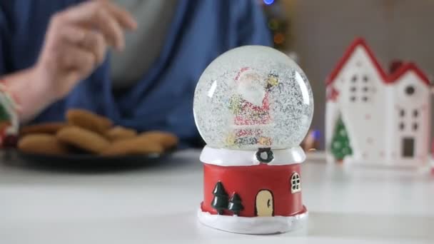 Скляна кулька зі снігом, на задньому плані чоловік їсть домашнє смачне різдвяне імбирне печиво зі склянкою — стокове відео