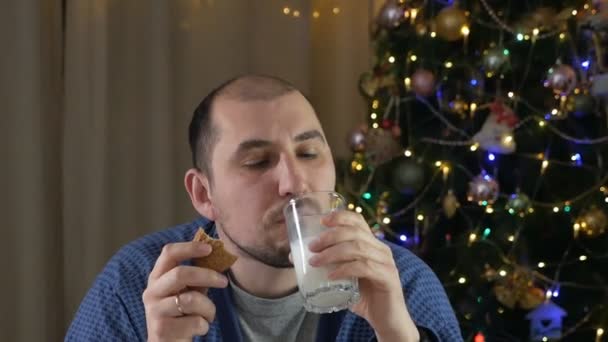 Um homem careca à noite em seu apartamento come deliciosos biscoitos de gengibre de Natal caseiros com um copo de leite perto de uma árvore de Natal com uma guirlanda. — Vídeo de Stock