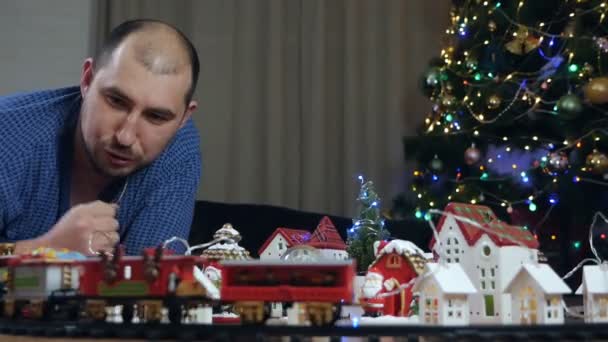 Ein Mann im Gewand spielt auf dem Boden mit einer Spielzeugeisenbahn und Eisenbahnen. Spielzeugweihnachtsstadt am Weihnachtsbaum — Stockvideo