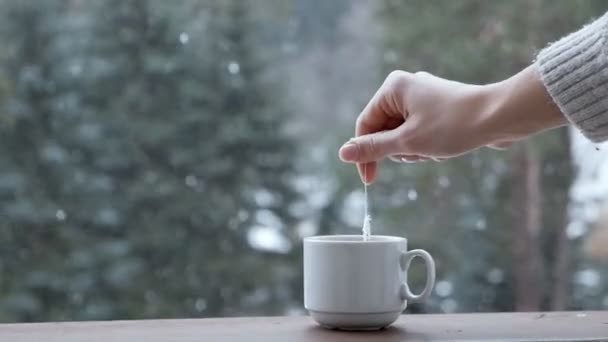 Ręka kładzie paczkę herbaty do białego kubka na parapecie na tle śnieżnej pogody. Śnieg, zima, ciepło, komfort — Wideo stockowe