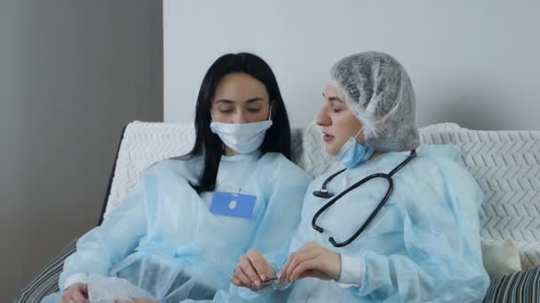 Jeunes femmes médecins en uniforme bleu portant un masque s'asseoir et discuter sur le canapé après un travail acharné — Video