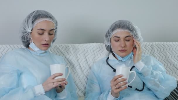 Müde, traurige Ärztinnen, die Kaffee auf der Couch trinken. Frustrierte Ärzte ruhen sich aus, machen eine Pause, nippen an einem Drink. Coronavirus. Entspannen Sie sich bei der Arbeit. COVID-19 — Stockvideo