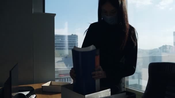 Egy nő, aki elvesztette az állását orvosi maszkban. Egy lány papírt tesz egy dobozba. Kirúgtak egy nőt a munkahelyéről. A vállalkozás csődje. Munkaveszteség — Stock videók