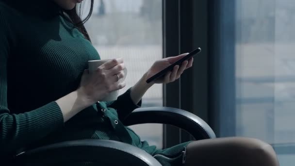 Eine Frau trinkt Tee und blickt im Büro eines Business Centers in der Nähe eines großen Panoramafensters auf das Telefon. — Stockvideo
