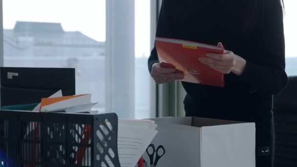 Молодая работница стоит за столом в офисной комнате. Упаковывает свои рабочие вещи в картонную коробку из-за увольнения. грустно и обидно.. — стоковое видео