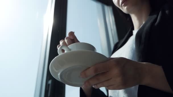 Jeune femme debout près de la fenêtre agitant une tasse de café dans le bureau — Video