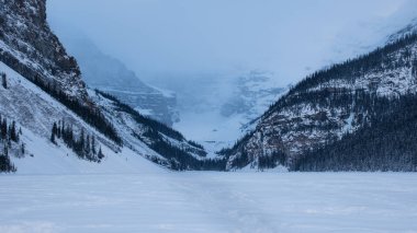 Donmuş bir gölde karlı bir yol Kanada kayalık dağ manzarası ve sisin içindeki buzullu dağ zirvesi. Alacakaranlıkta dondurucu soğuk ve şiddetli kar yağışı. Louise Gölü, Alberta, Kanada 'da görkemli ve gizemli dağ sıraları