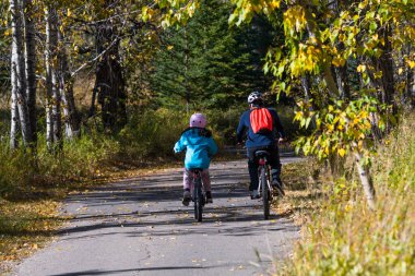 Ebeveyn ve çocuk parkta bisiklet sürüyor. Baba ve kızı uzun bir sonbahar haftasonunda bisiklete biniyorlar. Aile aktivitesi ve eğlenceli bir yaşam tarzı.