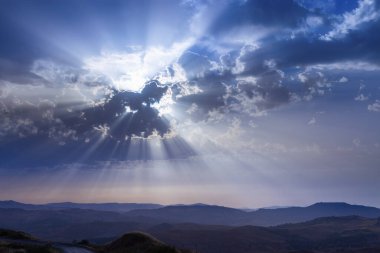 Güneş ışınları bulutları delip geçiyor. Tepenin ve dağların üzerindeki mavi gökyüzü. Dramatik bir gökyüzü. Güzel kavramsal meditasyon arkaplanı.