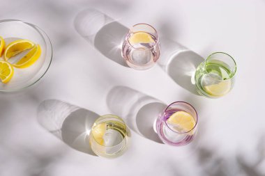 Güneşli bir günde beyaz arka planda bir dilim limonla renkli cam bardaklarda doğal su içmek. Sert gölgelerle bezenmiş yaz ayları. Üst görünüm.