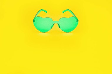Minimum moda fotoğrafçılığı kalp şeklindeki gözlüklerle parlak sarı kağıt arka planda. Yeşil modern güneş gözlüğü. Son moda yaz konsepti. Boşluğu kopyala.