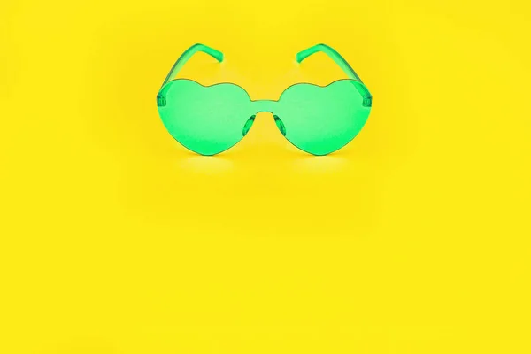 Minimal style fashion photography with heart shaped glasses on bright yellow paper background. Green modern sunglasses. Trendly summer concept. Copy space.