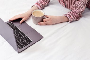 Laptop kullanan ve evde çalışan bir kız. Yatakta oturuyor ve pembe çizgili pijamalar giyiyor. Bir fincan kahve tutan ve bilgisayar ekranına bakan bir kadın..