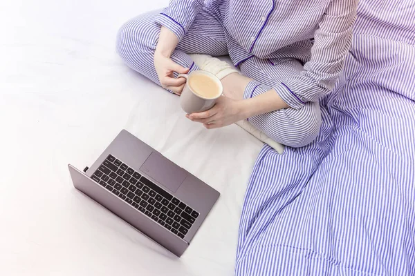 Laptop kullanan ve evde çalışan bir kız. O, yatakta oturuyor ve mavi çizgili pijamalar giyiyor. Bir fincan kahve tutan ve bilgisayar ekranına bakan bir kadın..