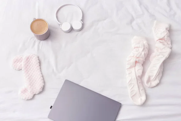 Work at home, rays of morning sun and randomly scattered things on bed. Flat lay with laptop, warm socks, cup of coffee, wireless headphones, mask for sleeping on bed sheet.