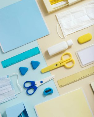 Okul malzemelerinin planı, defterler, kalemler, tıbbi maske ve el dezenfektanı kağıt arka planda, mavi ve sarı renkli. Yeni normal okula dönüş konsepti. Üst görünüm.