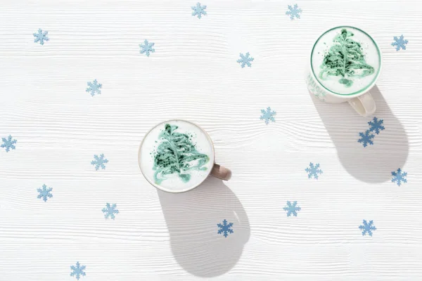 Latte winter coffee with New year tree art in big mug on white table with blue paper decor snowflakes. Background with hot beverage. Flat lay and hard shadows.