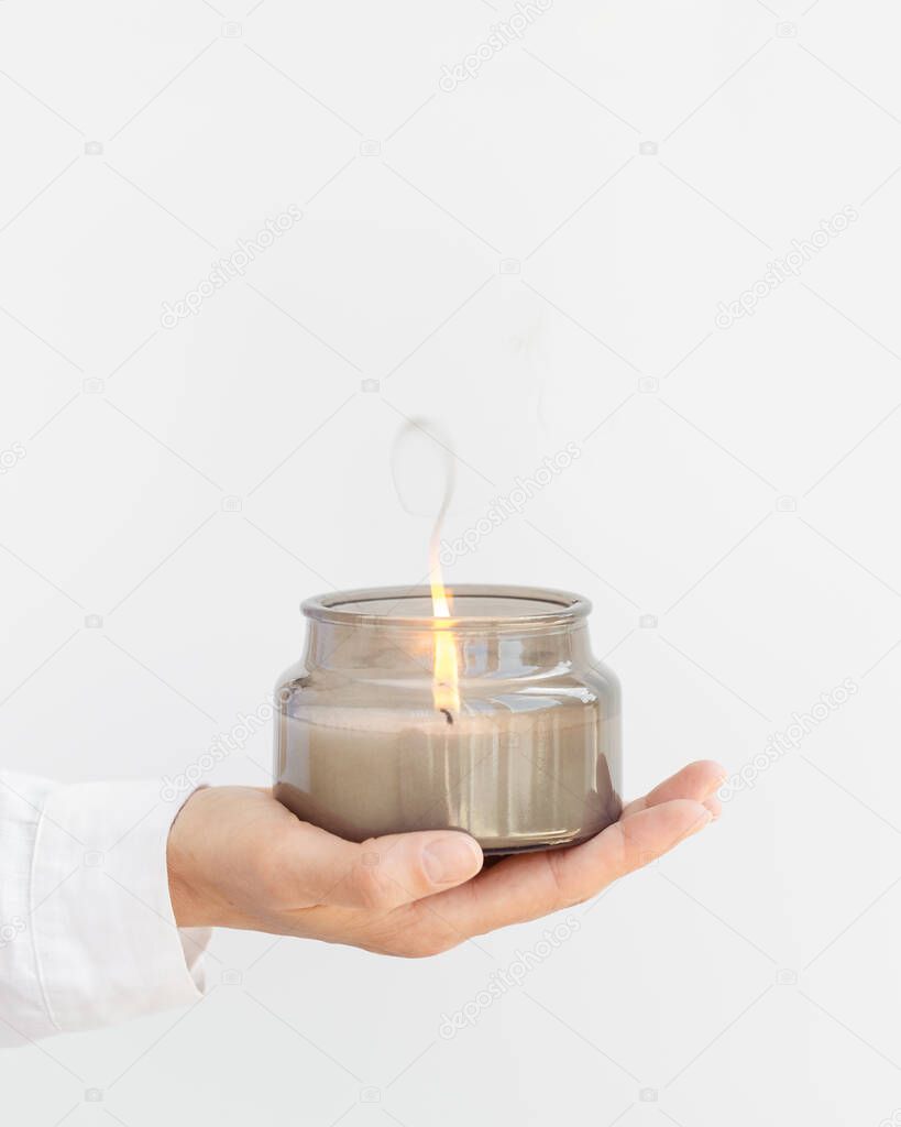 Woman hand holds lit scented candle in gray glass jar with natural ingredients on white background. Wellness and physical, emotional health concept.