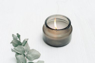 Cam kavanozda, beyaz ahşap arka planda, okaliptüs dallarıyla birlikte doğal malzemelerle yanan mum kokulu.