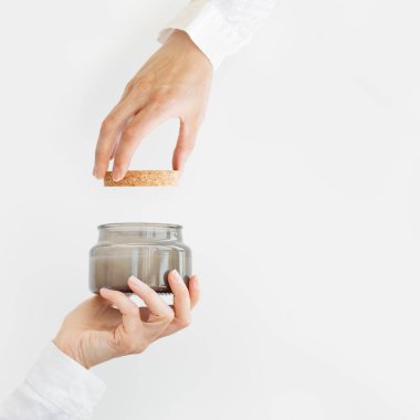 Kadın eli kapağı tutuyor ve cam kavanozdaki kokulu mumu beyaz arka planda doğal malzemelerle kapatıyor. Sağlık ve fiziksel, duygusal sağlık konsepti.