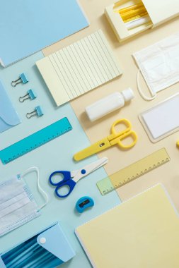 Okul malzemelerinin planı, defterler, kalemler, tıbbi maske ve el dezenfektanı kağıt arka planda, mavi ve sarı renkli. Yeni normal okula dönüş konsepti. Üst görünüm.