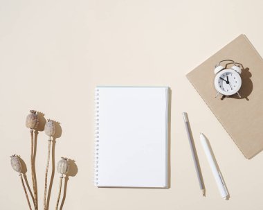 Boş sayfaları ve küçük saati olan açık defterin üst görüntüsü, masada kuru haşhaş, sonbahar konsepti. Yaratıcı çalışma alanı nötr bej tonları.