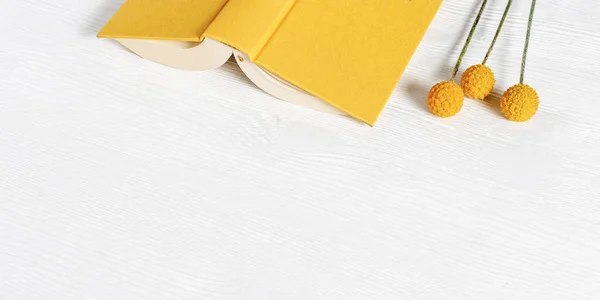 Open book in yellow cover and decorative flowers on white wooden table. Concept of reading books and time without gadgets. Top view with copy space.