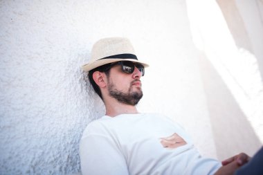 Hasır şapkalı ve güneş gözlüklü genç adam beyaz bir duvarın yanında şekerleme yapıyor.