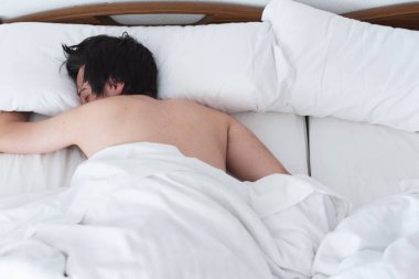Young man sleeping in a bed with white sheets, lying on his stomach. Time to wake up. Roam in bed.