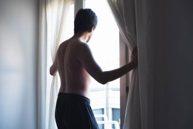 Young man naked from the waist up pulling some curtains and looking outside.