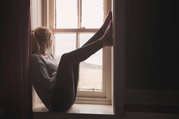A young woman stuck indoors sits on a window sill, gazing outside and longing to be free.