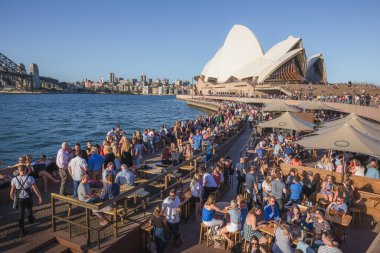 Opera binasına giden yoğun bir Sydney Limanı. Hem yerel halk hem de turistler işten sonra Opera Bar 'da içki içip sosyalleşiyorlar.. 