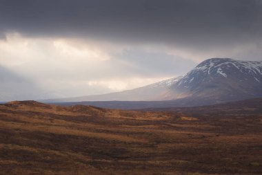 İskoç Highlands 'taki Glencoe' daki karlı dağ manzarasını aydınlatmak için alçak bulutların arasından dramatik bir güneş doğuyor..