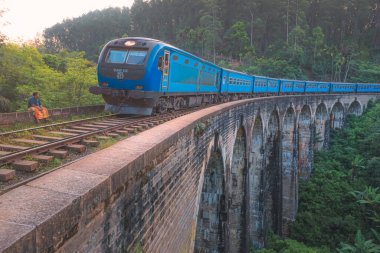 Sri Lankalı bir adam Demodara Dokuz Kemer Köprüsü viyadük demiryolunun yanında oturuyor. Mavi tren Sri Lanka 'nın orman tepesi ülkesinden geçerken..