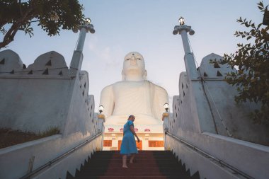 A young female caucasian tourist ascends the steps to the giant white Buddha statue at Sri Maha Bodhi Viharaya, a Buddhist temple at Bahirawakanda in Kandy, Sri Lanka. clipart
