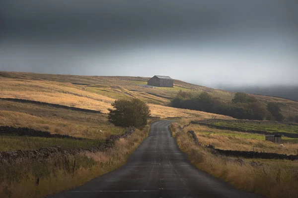 An old stone barn and an empty country road through a moody English countryside rural landscape in the North Pennines AONB, England UK.