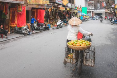Yerel bir Vietnamlı sokak satıcısı, geleneksel bir konik non la veya çiçek şapkalı Hanoi şehir sokaklarında bisikletinin arkasından meyve satıyor..