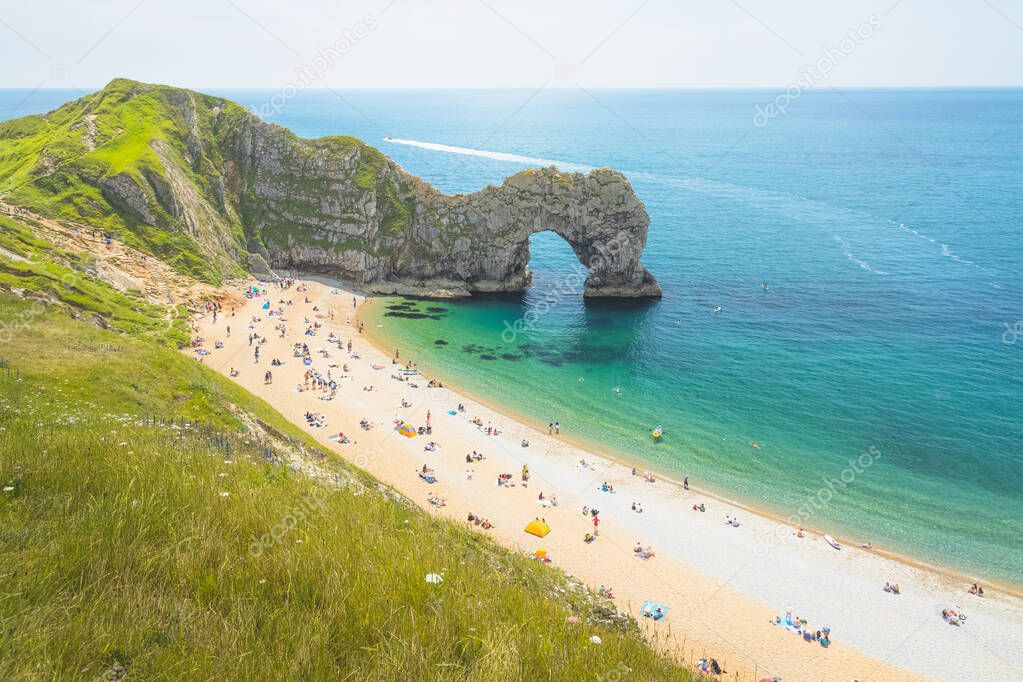 Tourists at the beach at the iconic sea arch Durdle Door on a sunny summer day on the Jurassic Coast in Dorset, UK.