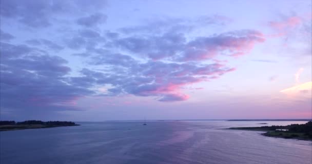 Letecký záběr letí přes klidný přístav Atlantského oceánu s jednou plachetnicí během barevného růžového a fialového západu slunce u pobřeží Maine.
