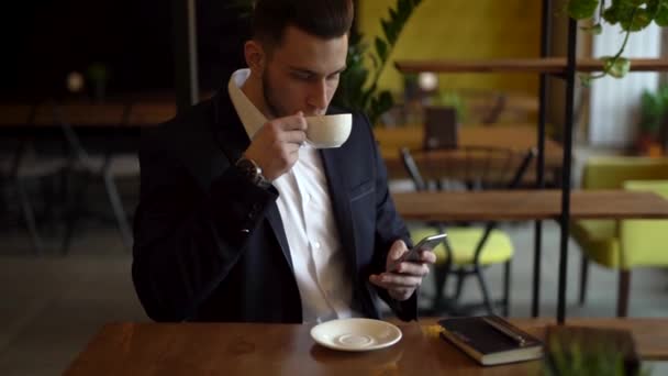 一个穿西装的年轻人正坐在咖啡馆里 男人有胡子 他在喝咖啡和咀嚼他的手机 桌子上还有笔记本和钢笔 — 图库视频影像