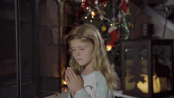 A small girl child sits by the fireplace in a festively decorated home and prays.