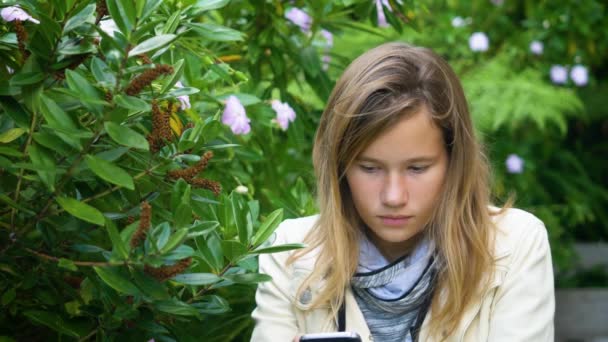 魅力的なティーンの女の子は庭に座っているスマートフォンでソーシャルメディアのメッセージをチェックします — ストック動画