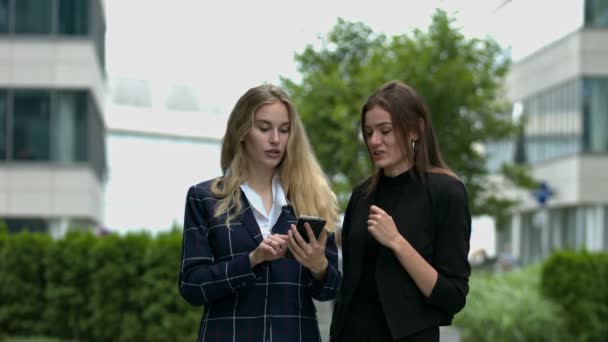 Két okosan öltözött nő néz egy mobilkészüléket, miközben egy modern városi környezetben beszélgetnek.