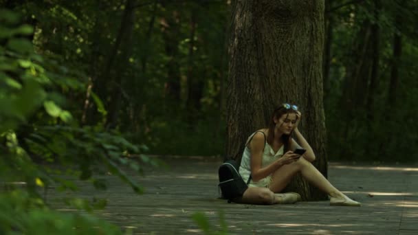 Chica Aburrida Sentada Contra Desplazamiento Del Árbol Teléfono Inteligente — Vídeo de stock