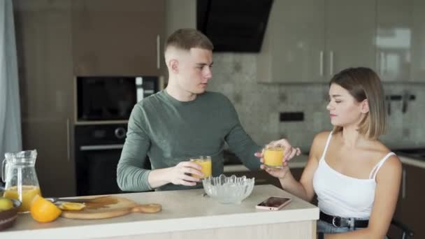 素敵なカップルはキッチンに座ってオレンジジュースを一緒に飲んでいます ペアは愛と抱擁にある — ストック動画