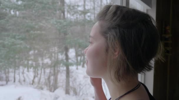 年轻快乐的女孩看着雪从窗户滑落 慢动作 — 图库视频影像